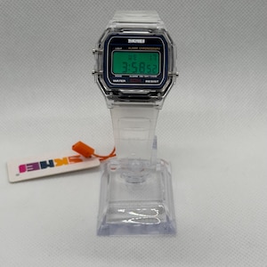 K&ouml;nnte beinhalten: Transparente Digitaluhr mit quadratischem Zifferblatt und transparentem Armband. Das Zifferblatt zeigt die Uhrzeit, den Tag und das Datum in Gr&uuml;n auf dunkelblauem Hintergrund an. Die Marke "SKMEI" ist sichtbar. Die Uhr steht auf einem transparenten St&auml;nder.