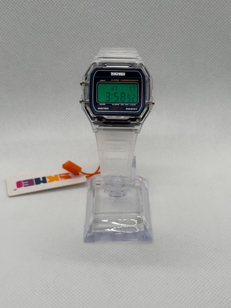 K&ouml;nnte beinhalten: Transparente Digitaluhr mit quadratischem Zifferblatt und transparentem Armband. Das Zifferblatt zeigt die Uhrzeit, den Tag und das Datum in Gr&uuml;n auf dunkelblauem Hintergrund an. Die Marke "SKMEI" ist sichtbar. Die Uhr steht auf einem transparenten St&auml;nder.