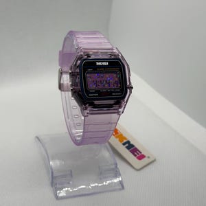 Puede incluir: Un reloj digital transparente de color lavanda con una esfera negra y la marca "SKMEI". El reloj tiene una esfera rectangular y una correa lavanda a juego. El reloj se exhibe en un soporte de pl&aacute;stico transparente.