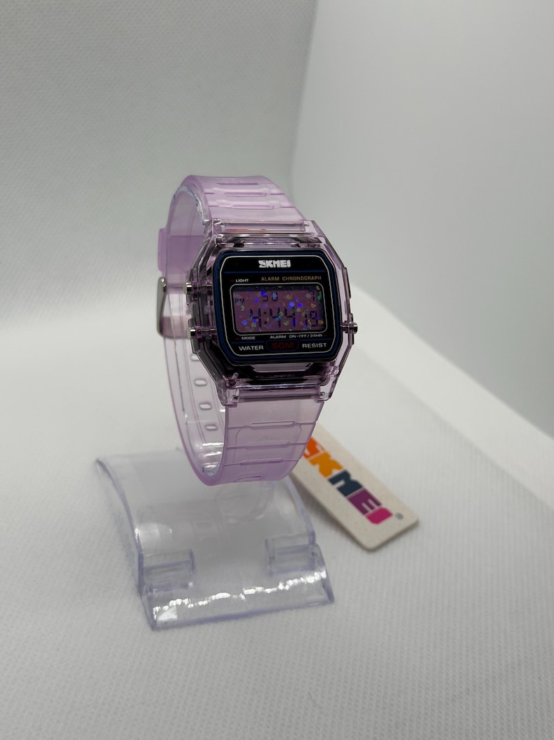 Puede incluir: Un reloj digital transparente de color lavanda con una esfera negra y la marca "SKMEI". El reloj tiene una esfera rectangular y una correa lavanda a juego. El reloj se exhibe en un soporte de pl&aacute;stico transparente.