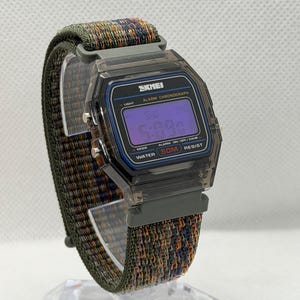 May include: A digital wristwatch with a translucent dark gray rectangular case and a purple digital display. The watch face features the text SKMEI, ALARM CHRONOGRAPH, and WATER 50M RESIST. It has a textured, multi-colored woven fabric strap.