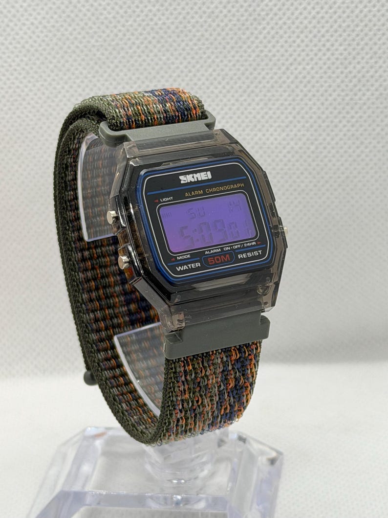 May include: A digital wristwatch with a translucent dark gray rectangular case and a purple digital display. The watch face features the text SKMEI, ALARM CHRONOGRAPH, and WATER 50M RESIST. It has a textured, multi-colored woven fabric strap.