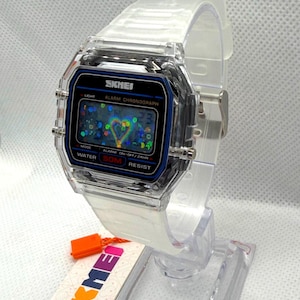 May include: A digital wristwatch with a transparent clear plastic case and matching strap. The watch face features a black border with blue accents, displaying the text SKMEI, ALARM CHRONOGRAPH, and WATER 50M RESIST. The screen shows a heart graphic.