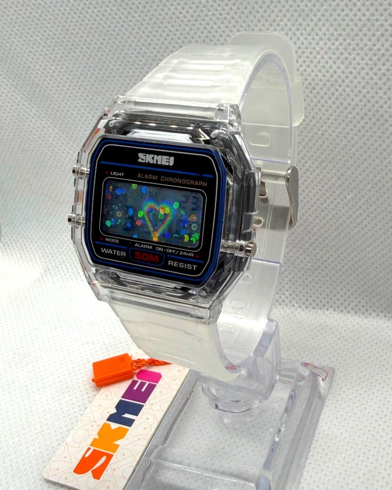 May include: A digital wristwatch with a transparent clear plastic case and matching strap. The watch face features a black border with blue accents, displaying the text SKMEI, ALARM CHRONOGRAPH, and WATER 50M RESIST. The screen shows a heart graphic.