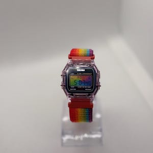 May include: A digital watch with a rainbow-colored strap. The watch face displays the time, with the brand name "SKMEI" visible. The strap is a vibrant gradient of colors, and the watch case is a transparent pink.