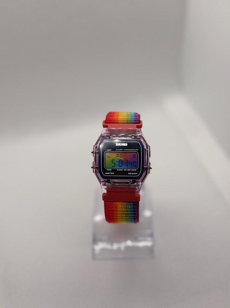 May include: A digital watch with a rainbow-colored strap. The watch face displays the time, with the brand name "SKMEI" visible. The strap is a vibrant gradient of colors, and the watch case is a transparent pink.