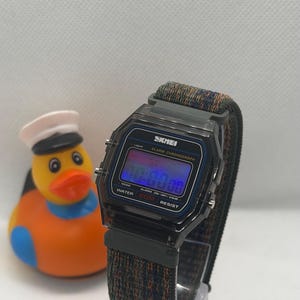 May include: A digital wristwatch with a translucent black rectangular case and a dark, multi-colored woven fabric strap. The watch face displays the brand name SKMEI, Alarm Chronograph, and Water 50M Resist. A yellow rubber duck with a white hat is nearby.