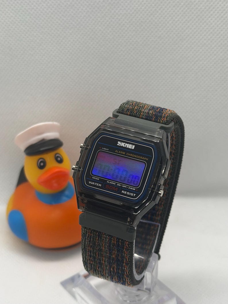 May include: A digital wristwatch with a translucent black rectangular case and a dark, multi-colored woven fabric strap. The watch face displays the brand name SKMEI, Alarm Chronograph, and Water 50M Resist. A yellow rubber duck with a white hat is nearby.