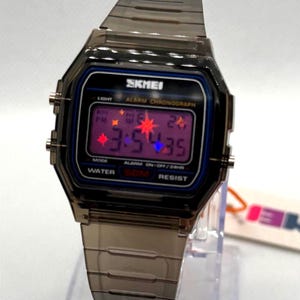 May include: A digital wristwatch with a translucent dark grey plastic case and matching link band. The rectangular face features the brand name SKMEI, a purple digital display showing 3:54:35, and text including ALARM CHRONOGRAPH and WATER 50M RESIST.