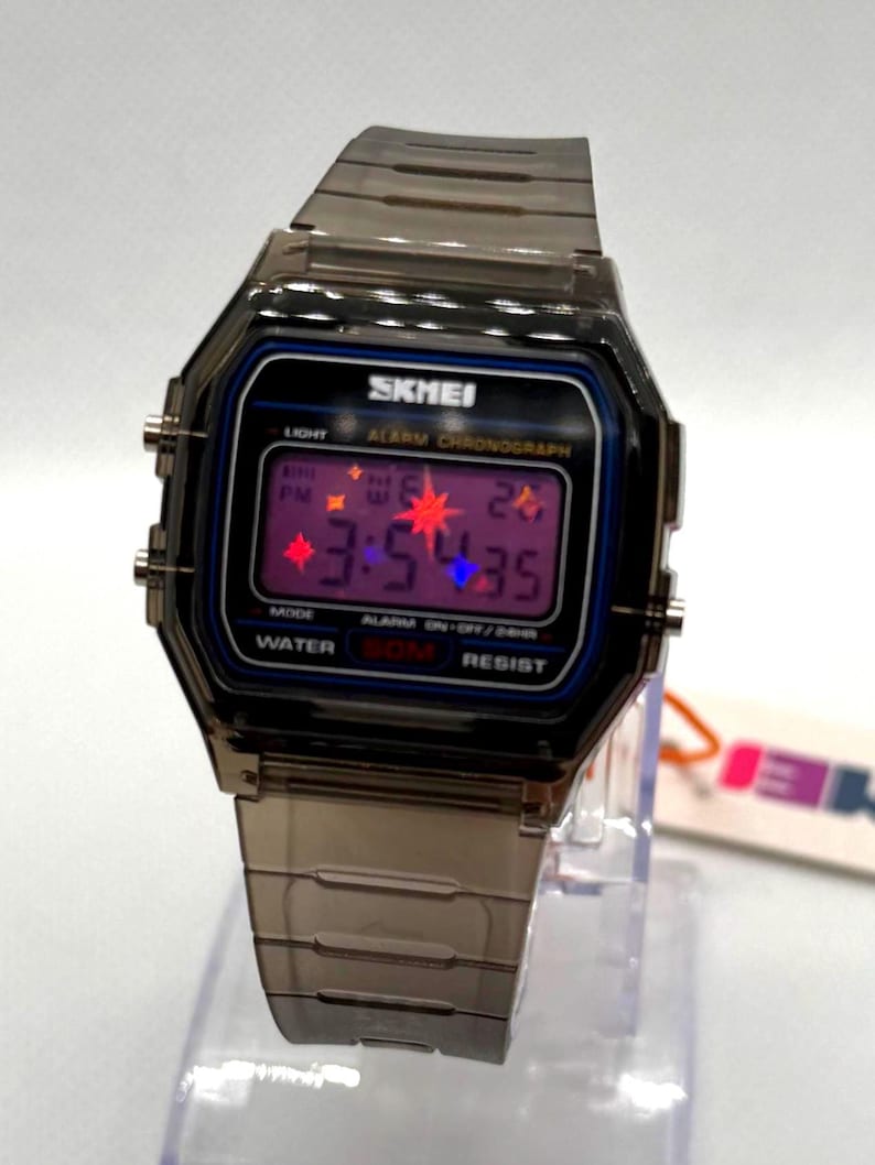May include: A digital wristwatch with a translucent dark grey plastic case and matching link band. The rectangular face features the brand name SKMEI, a purple digital display showing 3:54:35, and text including ALARM CHRONOGRAPH and WATER 50M RESIST.