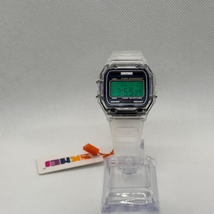 K&ouml;nnte beinhalten: Transparente Digitaluhr mit rechteckigem Zifferblatt und transparentem Armband. Die Uhr zeigt die Uhrzeit 3:59:09 auf einem gr&uuml;nen Bildschirm an. Die Uhr steht auf einem transparenten Pr&auml;sentationsst&auml;nder, und ein Etikett mit dem Markennamen ist angebracht.