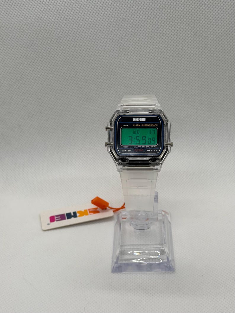 K&ouml;nnte beinhalten: Transparente Digitaluhr mit rechteckigem Zifferblatt und transparentem Armband. Die Uhr zeigt die Uhrzeit 3:59:09 auf einem gr&uuml;nen Bildschirm an. Die Uhr steht auf einem transparenten Pr&auml;sentationsst&auml;nder, und ein Etikett mit dem Markennamen ist angebracht.