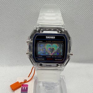 May include: A digital wristwatch with a clear, transparent plastic case and matching translucent strap. The rectangular black watch face displays the brand name SKMEI, the text ALARM CHRONOGRAPH, and WATER 50M RESIST, featuring a holographic heart graphic.