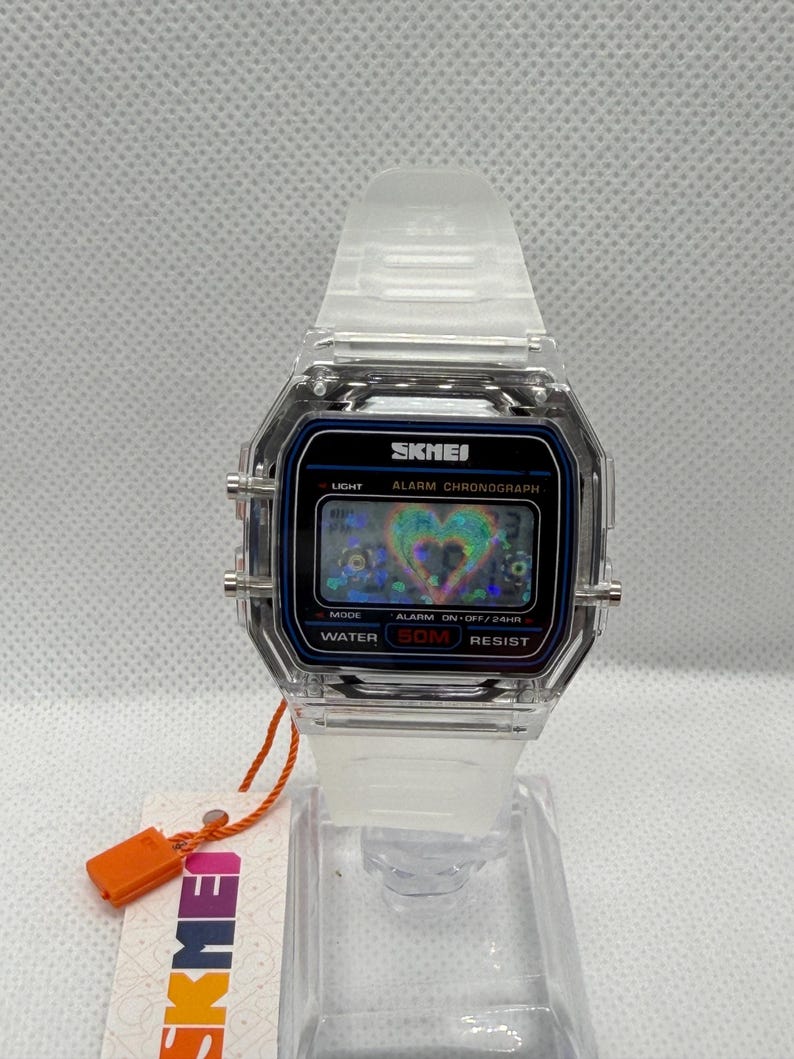May include: A digital wristwatch with a clear, transparent plastic case and matching translucent strap. The rectangular black watch face displays the brand name SKMEI, the text ALARM CHRONOGRAPH, and WATER 50M RESIST, featuring a holographic heart graphic.