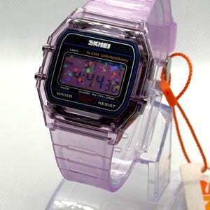 Puede incluir: Un reloj digital lavanda transparente con una esfera negra y la marca "SKMEI". El reloj muestra la hora y tiene una correa lavanda a juego. El reloj es resistente al agua y tiene funciones de alarma y cronógrafo.