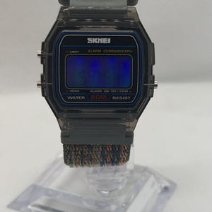 May include: A digital wristwatch with a translucent dark gray rectangular case and a blue-tinted display screen. The watch face features the text SKMEI, ALARM CHRONOGRAPH, and WATER 50M RESIST. It includes a textured fabric strap in mixed gray and orange tones.