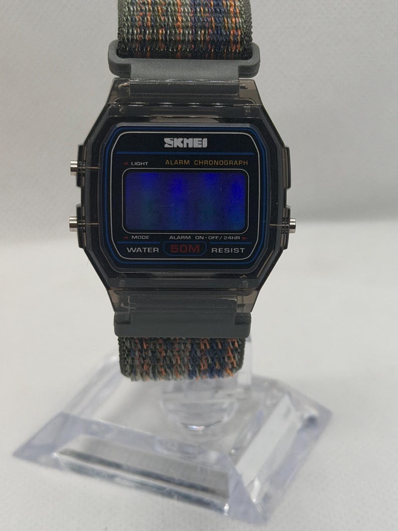 May include: A digital wristwatch with a translucent dark gray rectangular case and a blue-tinted display screen. The watch face features the text SKMEI, ALARM CHRONOGRAPH, and WATER 50M RESIST. It includes a textured fabric strap in mixed gray and orange tones.