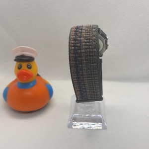 May include: A digital watch with a textured, multi-colored woven strap in shades of grey, blue, and orange, mounted on a clear stand, positioned next to a yellow rubber duck wearing a white sailor hat with a black brim and a blue neck band.