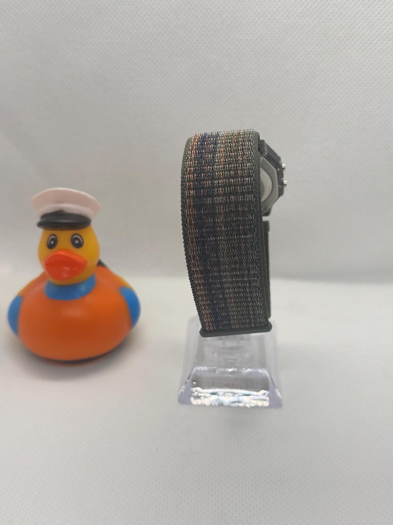 May include: A digital watch with a textured, multi-colored woven strap in shades of grey, blue, and orange, mounted on a clear stand, positioned next to a yellow rubber duck wearing a white sailor hat with a black brim and a blue neck band.