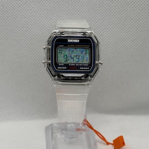May include: A digital wristwatch with a transparent clear plastic case and matching white strap. The rectangular watch face features a black border, a digital display showing the time and date, and the text SKMEI, ALARM CHRONOGRAPH, and WATER 50M RESIST.
