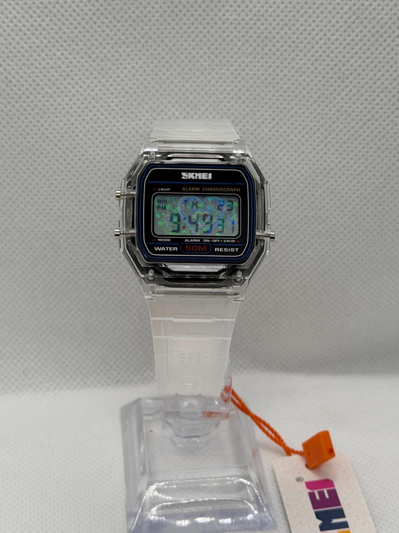 May include: A digital wristwatch with a transparent clear plastic case and matching white strap. The rectangular watch face features a black border, a digital display showing the time and date, and the text SKMEI, ALARM CHRONOGRAPH, and WATER 50M RESIST.