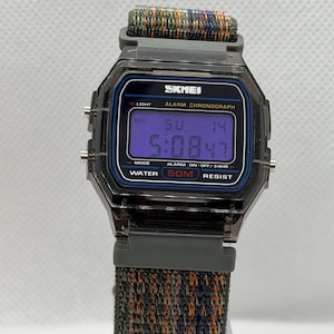 May include: A digital wristwatch with a translucent dark gray case and a multi-colored woven fabric strap. The watch face displays the brand name SKMEI, the text ALARM CHRONOGRAPH, and WATER 50M RESIST. The digital screen shows the time 5:08 and date SU 14.