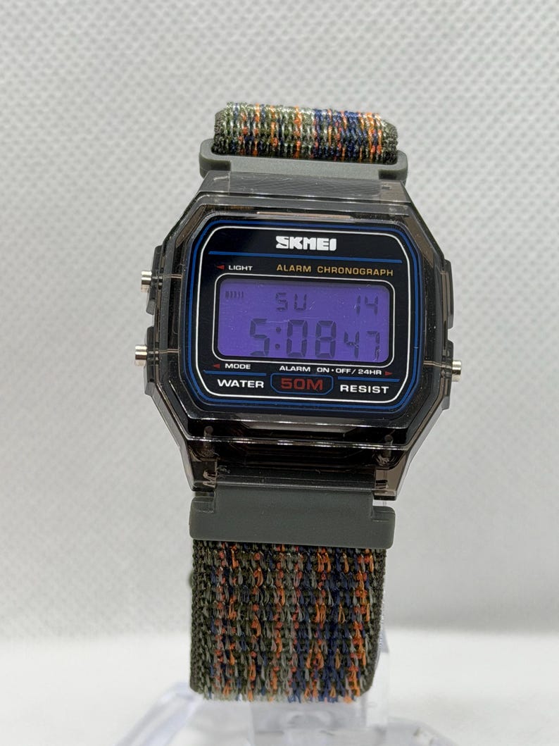 May include: A digital wristwatch with a translucent dark gray case and a multi-colored woven fabric strap. The watch face displays the brand name SKMEI, the text ALARM CHRONOGRAPH, and WATER 50M RESIST. The digital screen shows the time 5:08 and date SU 14.
