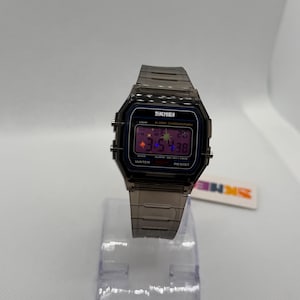 May include: A digital wristwatch with a translucent dark grey resin case and matching link strap. The rectangular face features a black border, a digital display showing 3:54:38, and the text SKMEI, ALARM CHRONOGRAPH, and WATER RESIST.