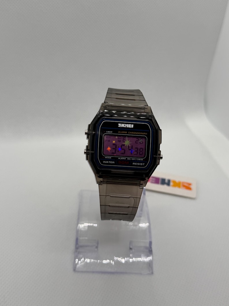 May include: A digital wristwatch with a translucent dark grey resin case and matching link strap. The rectangular face features a black border, a digital display showing 3:54:38, and the text SKMEI, ALARM CHRONOGRAPH, and WATER RESIST.