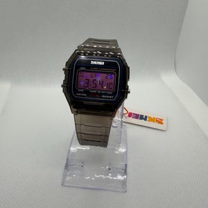 May include: A digital wristwatch with a translucent dark grey plastic case and matching link strap. The rectangular face features the text SKMEI, ALARM CHRONOGRAPH, and WATER RESIST. The digital display shows the time 3:54 and the date.