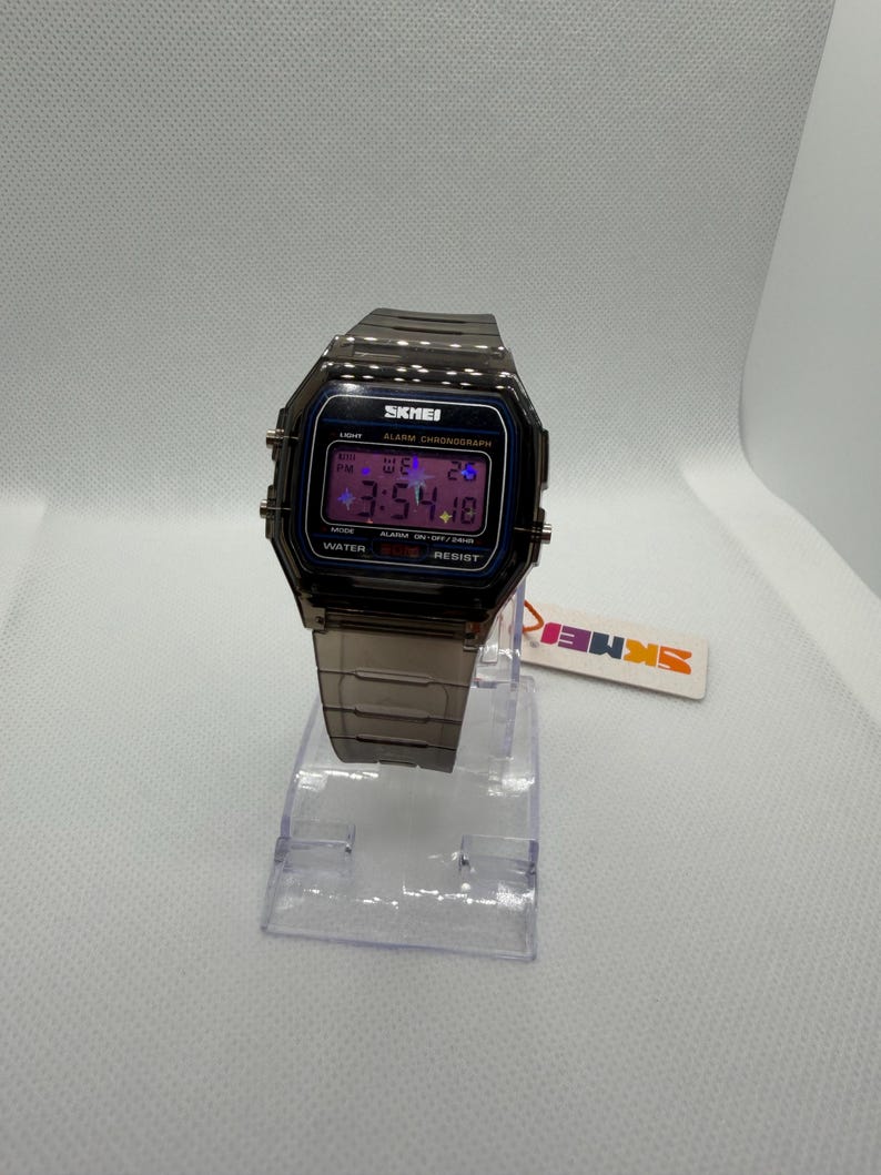 May include: A digital wristwatch with a translucent dark grey plastic case and matching link strap. The rectangular face features the text SKMEI, ALARM CHRONOGRAPH, and WATER RESIST. The digital display shows the time 3:54 and the date.