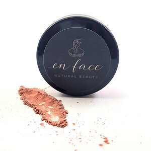Może przedstawiać: Czarny słoiczek z pudrem z okrągłą etykietą z motywem baletnicy i napisem "en face NATURAL BEAUTY" w ozdobnej czcionce. Obok słoiczka na białej powierzchni znajduje się smuga różowego pudru.