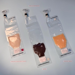 以下が含まれることがあります： 「Pineapple-Grapefruit Drink」、「Coffee」、「Orange Drink」とラベルされた粉末飲料が入った3つの透明なポーチ。ポーチにはノズルと小さなラベルが付いています。「Apollo Beverages」の文字が上部にあります。