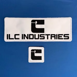 May include: A rectangular white embroidered patch featuring a black logo and the text ILC INDUSTRIES in black, accompanied by a smaller square white patch with the same black logo. Both patches have a white border and a clean, flat design.