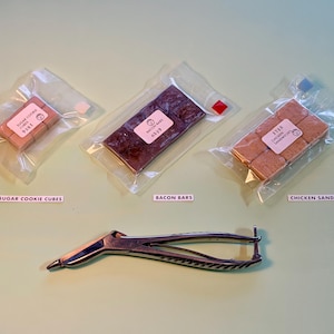 Peut inclure: Plusieurs aliments emballés, notamment des cubes de biscuits au sucre, des barres de bacon et des sandwichs au poulet, sont présentés. Un outil en métal argenté est également présent. Les articles sont sur une surface vert clair.