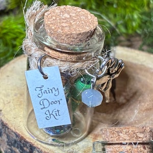 May include: A small glass jar with a cork stopper and a twine wrap, labeled "Fairy Door Kit." The jar contains a green bead, a blue gem, and glitter. A silver charm and a small tag are attached.