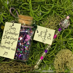 Könnte beinhalten: Eine kleine Glasflasche gefüllt mit lila Glitzer und einem Feenstab. Die Flasche hat einen Korken und ein Etikett mit der Aufschrift "Fairy Godmother Dust brings bouts of happiness and joy." Der Zauberstab ist mit Perlen und einem Kristall verziert.