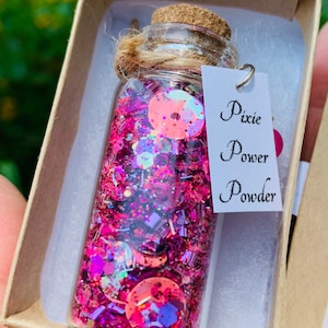 Könnte beinhalten: Eine kleine Glasflasche gefüllt mit rosa und lila Glitzer, Pailletten und schillernden Formen. Die Flasche hat einen Korken und ist mit Bindfaden gebunden. Ein kleines Etikett trägt die Aufschrift "Pixie Power Powder".