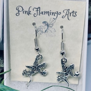 May include: Silver-toned fairy earrings with intricate details, displayed in a white box. The earrings feature a fairy design holding a small orb. The card behind the earrings reads "Pink Flamingo Arts".