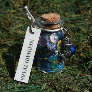 Puede incluir: Un pequeño frasco de vidrio lleno de lentejuelas y cuentas de colores, etiquetado como "MERMAID TEARS". El frasco tiene un tapón de corcho, un dije de metal y una etiqueta blanca. Contenido: lentejuelas azules, verdes e iridiscentes.