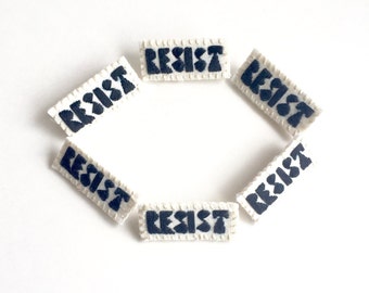 Hand embroidered brooch "Resist" black thread on bright cream muslin with sturdy cream felt backing An Astrid Endeavor activist