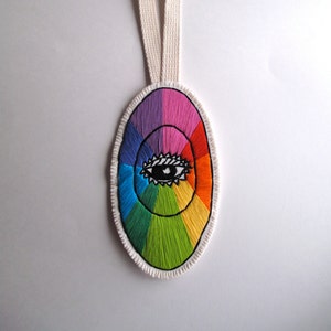 Könnte beinhalten: Ein bunter, gestickter ovaler Anhänger mit einem Regenbogen-Farbkreis und einem schwarzen Auge in der Mitte. Der Anhänger hat eine weiße Stoffschlaufe zum Aufhängen.