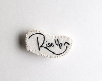 Rise up brooch hand embroidered with black writing on cream muslin with cream felt backing an astrid endeavor activist pin