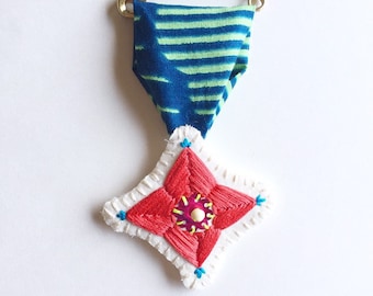 Hand embroidered talisman brooch with red blue yellow threads and pink sequin with blue and green wax fabric gold safety pin merit badge