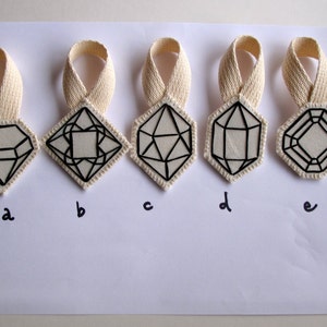 May include: Five geometric pendant designs with black outlines on a white background. Each pendant has a different internal design, with a cream-colored ribbon at the top. The pendants are labeled a, b, c, d, and e.