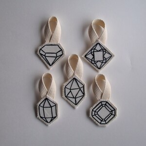 May include: Six white fabric ornaments with black geometric designs, each with a cream-colored loop for hanging. The shapes include diamonds, squares, and other polygonal forms. The ornaments are arranged on a white background.