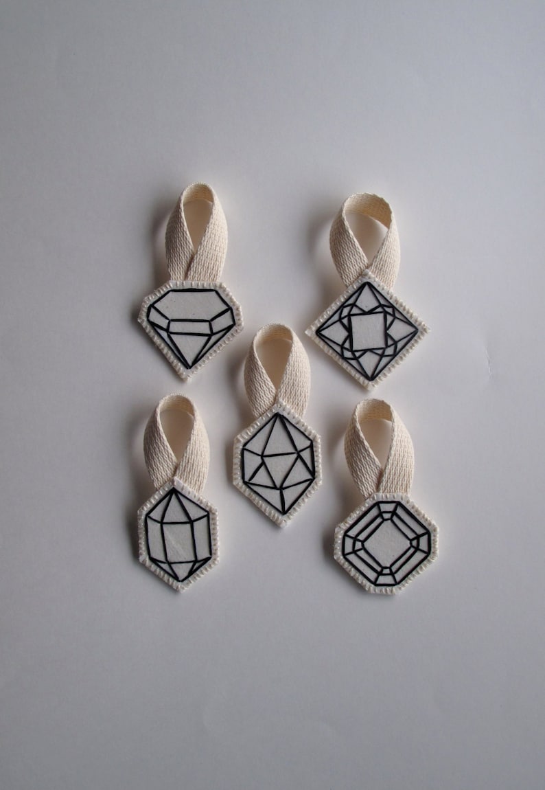 May include: Six white fabric ornaments with black geometric designs, each with a cream-colored loop for hanging. The shapes include diamonds, squares, and other polygonal forms. The ornaments are arranged on a white background.