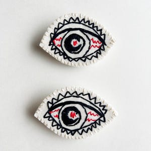 以下が含まれることがあります： 白い背景に黒と赤のステッチで刺繍された目のパッチが2つあります
