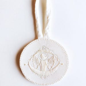 May include: White felt ornament with a white embroidered eye design. The ornament has a white fabric loop for hanging.