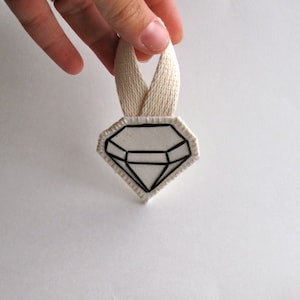 May include: A diamond-shaped, white and black graphic design with a cream-colored ribbon loop. The diamond design has a black outline and internal lines, creating a geometric pattern. The ribbon is held up by a hand.